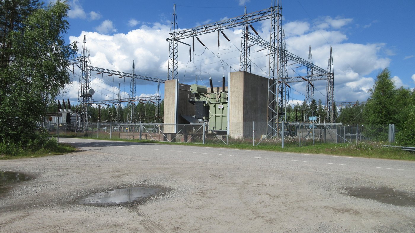 A large power transformer between concrete blast walls in a high voltage switchyard, with steel gantries and insulator strings overhead